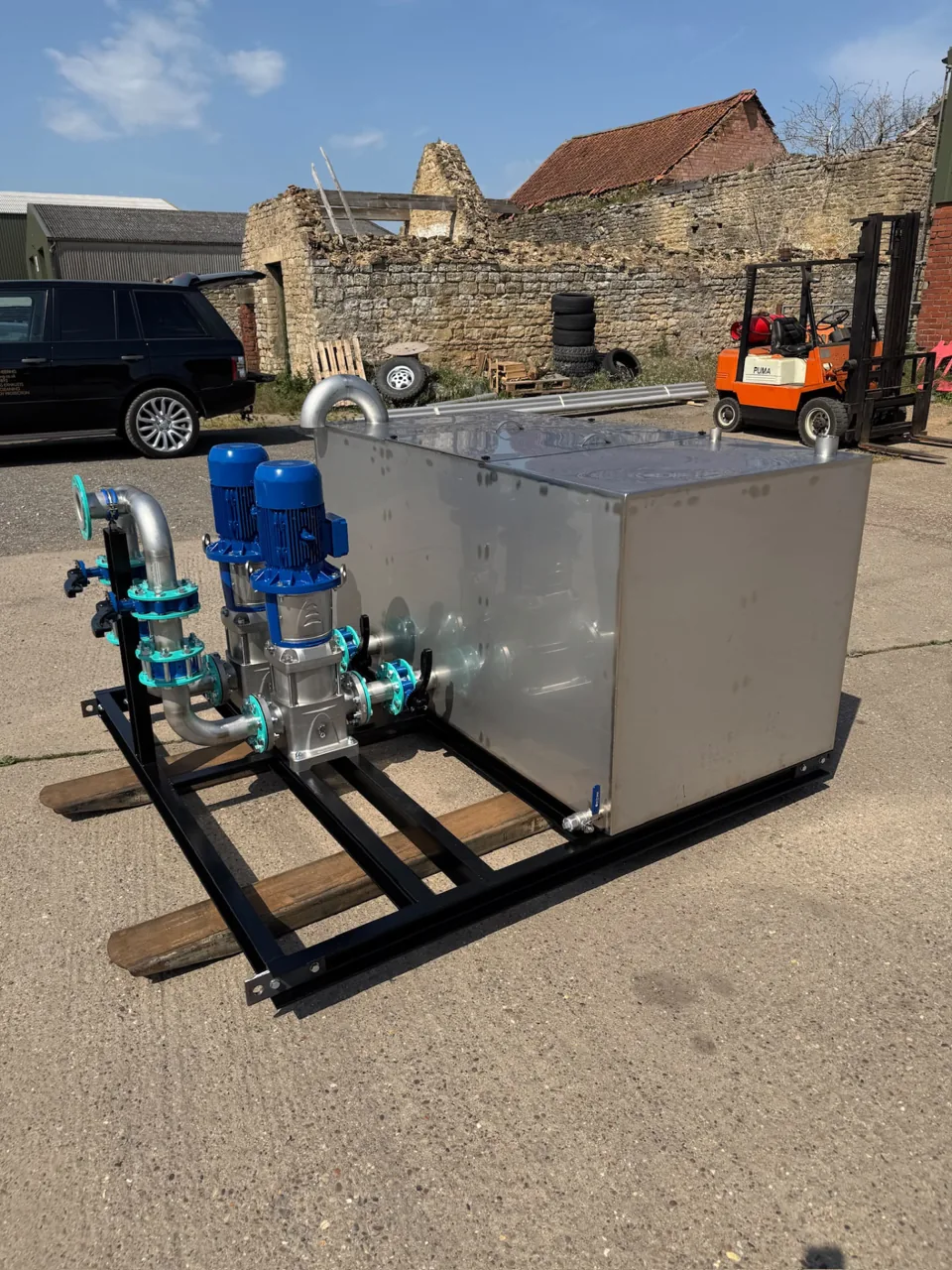 Rectangular stainless tank and pump assembly on a black skid in the workshop yard